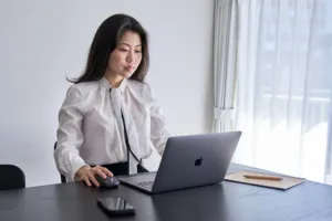 Business woman sitting at a dark wood desk using a laptop and mouse in a bright, minimalist office.