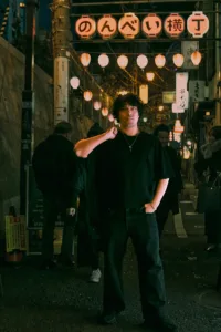 A professional night portrait of a man with curly hair standing in Nonbei Yokocho (Drunkard's Alley) in Shibuya, illuminated by warm street lanterns and professional lighting.