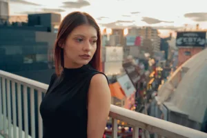 渋谷の街並みを背景に、ベランダでポーズをとる女性モデル。夕暮れ時のエモーショナルな光。