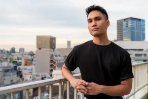A young entrepreneur in a black t-shirt posing on JG's Shibuya studio balcony with the Tokyo skyline in the background, showcasing urban portrait photography.