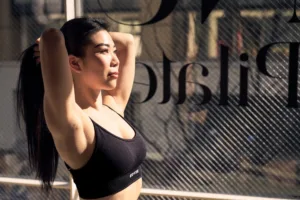 ハイコントラストなフィットネスブランド向けの写真。窓から差し込む温かい自然な逆光の中で、トレーニングウェアを着た女性が髪を整えている。背景には「Pilates」の文字が見える。