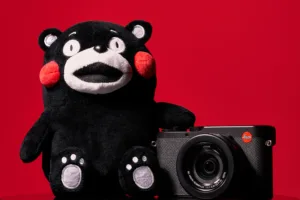 A black Kumamon plush mascot sitting next to a black Leica digital camera against a vibrant solid red background.