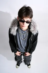 東京を拠点とするフォトグラファー、JGによるスタジオポートレート。Post Xeno（ポスト・ゼノ）のフェイクファー・フーデッドジャケット、有刺鉄線ネックレス、そしてダメージ加工のストリートウェアを身に纏ったモデルを、ハイアングルから捉えた一枚。