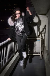 Model wearing a Post Xeno streetwear outfit on a Shibuya balcony at night, featuring a fur-lined hoodie, graphic t-shirt, and studded belt.