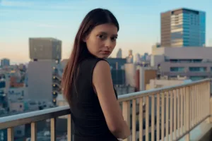 Young woman in black top posing on Tokyo balcony with cityscape background, urban portrait by JG Brasseur.
