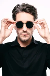 白のシームレス背景に、黒のレイバン・ラウンドサングラスとダークカラーのボタンダウンシャツを合わせたJGのハイコントラストなスタジオポートレート。明るくクリーンなライティングが、東京のファッションフォトグラフィーにふさわしい、プロフェッショナルかつモダンな美学を強調しています。