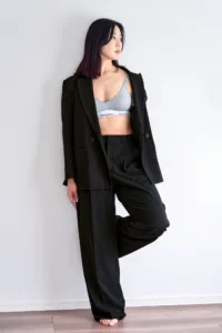 Fashion portrait of a woman in a black power suit and grey bralette against a white wall, by JG.