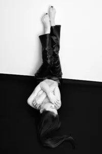 Black and white studio portrait of a woman in leather trousers lying with legs raised against a white wall.