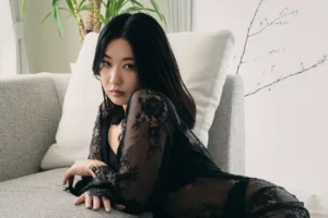 Natural light portrait of Rioka in a sheer black lace dress reclining on a grey sofa beside a cherry blossom branch at JG Studio in Tokyo.