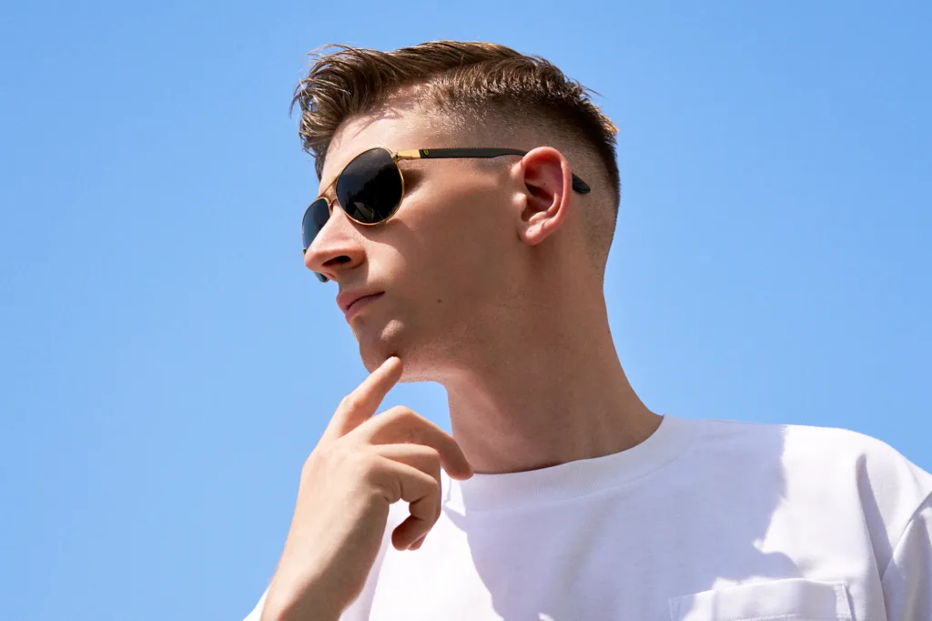 Low-angle outdoor portrait of a man wearing gold-framed sunglasses and a white pocket t-shirt against a clear blue sky, captured during an editorial session in Tokyo by JG.